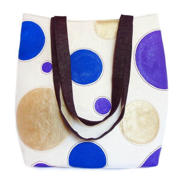 Tote bag featuring natural canvas hand painted with giant polka dots in gold, purple, and blue, paired with dark blue denim flecked with gold thread. 