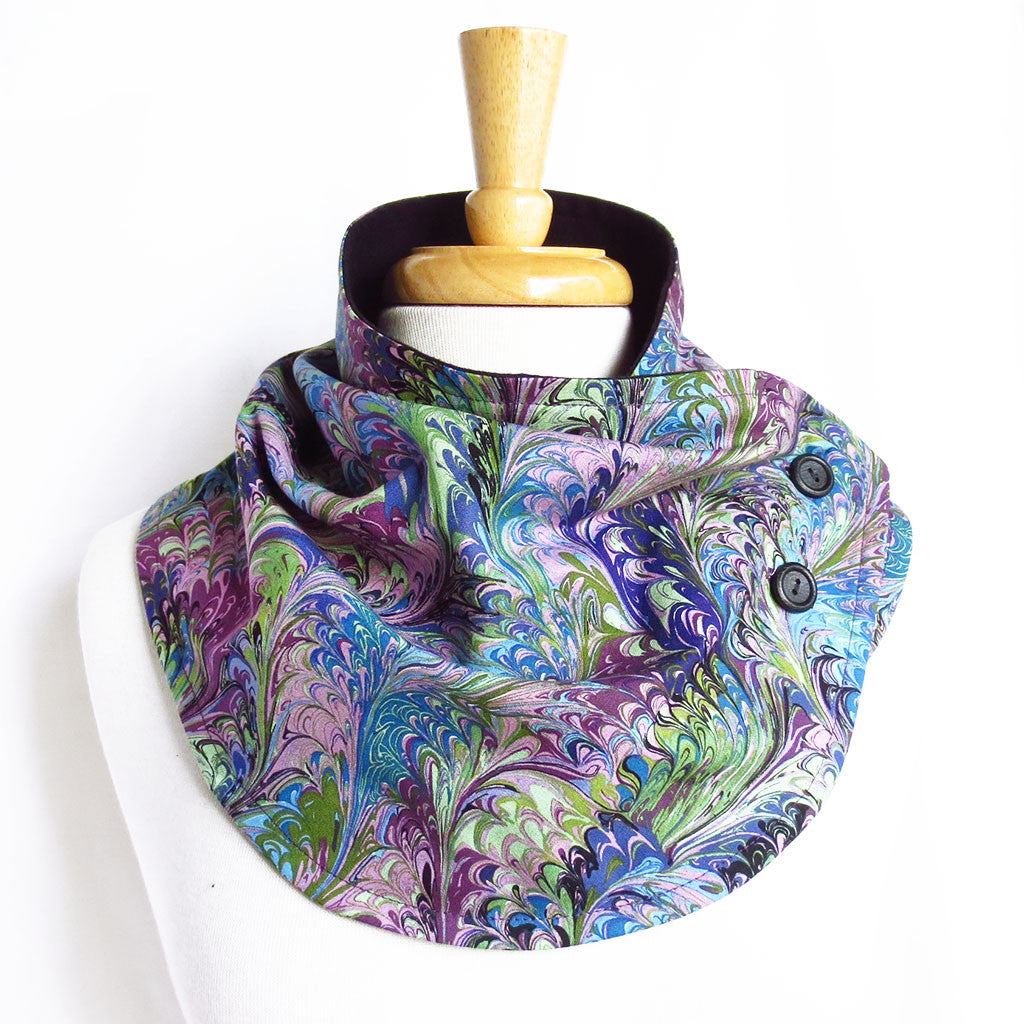 the natalie button scarf is in a classic book marble print in shades of purple, blue, and blue-green, with black hand painted buttons and black flannel lining